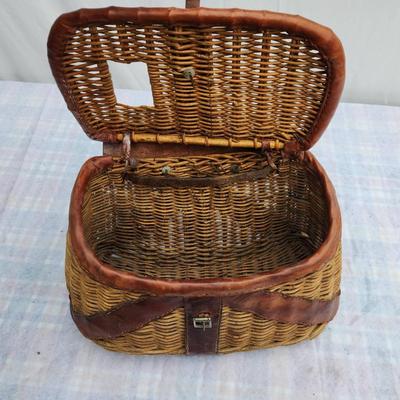 Sale Photo Thumbnail #147: Wicker fishing creel with leather trim and straps, featuring a lid with a slot for fish entry and a buckle closure mechanism for securing the contents. The creel is designed for outdoor fishing use, with carrying straps for convenience. In used condition,