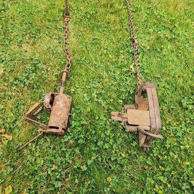 Sale Photo Thumbnail #30: Two vintage metal animal traps with attached chains. The set includes one cylindrical and one rectangular trap, each exhibiting rust and wear consistent with age and use. Both traps are intact with all moving parts. Each trap is connected to a chain that 