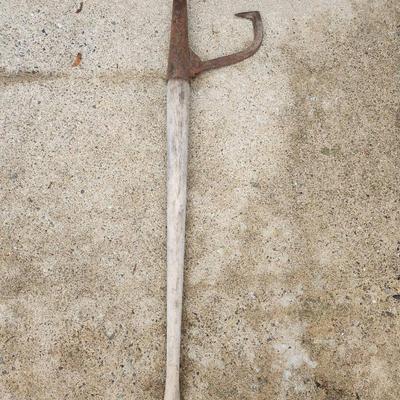 Sale Photo Thumbnail #194: A long-handled tool featuring a wooden shaft and a rusted iron head. The head includes one pointed end and one hooked element, likely for gripping or cutting applications. The item shows significant signs of rust and wear consistent with age and use.
