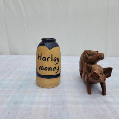 Sale Photo Thumbnail #221: A small ceramic jar labeled 'Harley money,' featuring a yellow body with a blue heart and band accent, alongside a wooden figurine of two pigs in a playful pose. The jar serves as a coin holder or decoration, while the wooden carving showcases detailed cr