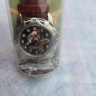 Sale Photo Thumbnail #231: Cal Ripken themed Sports Superstars watch, featuring a leather strap and a unique dial design. Comes in its original packaging, which showcases related artwork and details. The item appears to be labeled FSC: 24946-7, and it highlights the baseball supers