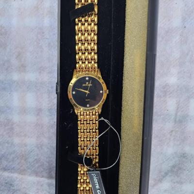 Sale Photo Thumbnail #233: Pierre Cardin gold-tone quartz watch featuring a black dial with a Roman numeral bezel and four crystals at the 3, 6, 9, and 12 o'clock positions. It comes with a multi-link gold-tone bracelet. Presented in its original box with tag attached. No visible d