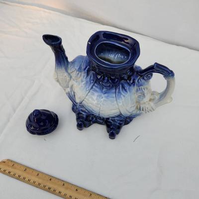 Sale Photo Thumbnail #238: Blue and white ceramic figural teapot, featuring a seated figure design. The teapot includes a removable lid and is hand-painted with gradient blue tones transitioning to white. Measuring tools shown suggest approximate size; exact dimensions are unstated