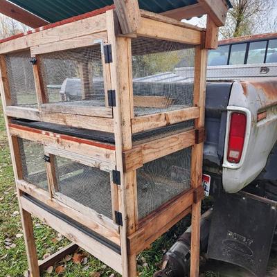 Sale Photo Thumbnail #45: A wooden quail cage with two levels, featuring wire mesh sides and bottoms for ventilation and waste management. The cage includes hinged front doors for easy access and a slanted green metal roof for weather protection. Supported by four wooden legs, it 
