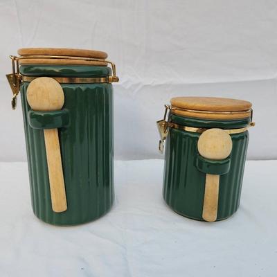 Sale Photo Thumbnail #276: Two ribbed ceramic canisters in a green finish, featuring hinged wooden lids and matching wooden spoons secured on the fronts. Each canister is fitted with a clasp closure to ensure a tight seal. These functional and decorative items are suitable for hold