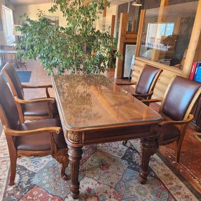 Sale Photo Thumbnail #18: Wooden dining table set measuring 6x3x2.6 feet, featuring a glass top and ornate carved detailing on the legs. Includes six chairs with padded seats and backs, upholstered in brown leather-like material, and armrests. Each chair displays detailed carvings