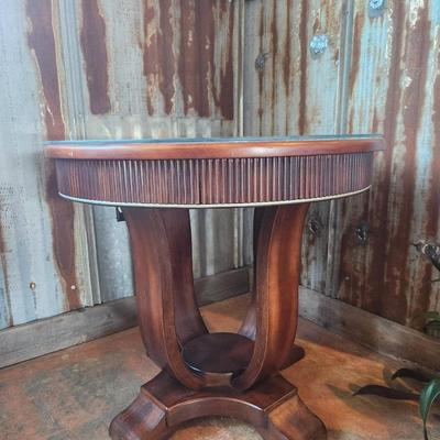 Sale Photo Thumbnail #27: Round accent table featuring a wood base with vertical detailing on the apron and a unique pedestal-style support. The tabletop is clear glass with beveled edges, secured atop the wooden frame. Appears to combine functional design with aesthetic detailing