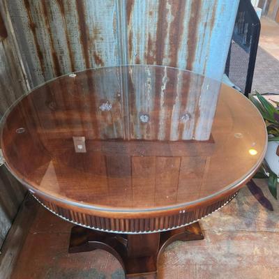 Sale Photo Thumbnail #28: Round accent table featuring a wood base with vertical detailing on the apron and a unique pedestal-style support. The tabletop is clear glass with beveled edges, secured atop the wooden frame. Appears to combine functional design with aesthetic detailing