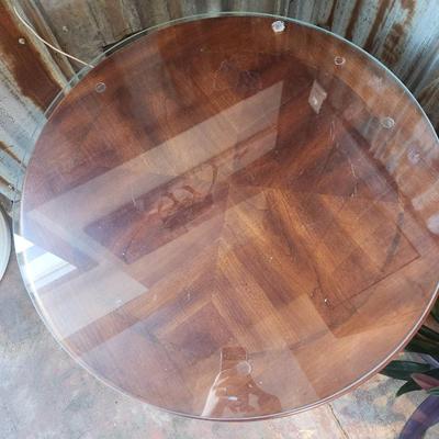 Sale Photo Thumbnail #29: Round accent table featuring a wood base with vertical detailing on the apron and a unique pedestal-style support. The tabletop is clear glass with beveled edges, secured atop the wooden frame. Appears to combine functional design with aesthetic detailing