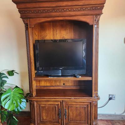 Sale Photo Thumbnail #35: A wooden entertainment armoire featuring carved details along the top and side columns. The unit includes an open space accommodating a flat-screen TV, a central shelf for media equipment, and two lower cabinet doors for additional storage. The armoire ha