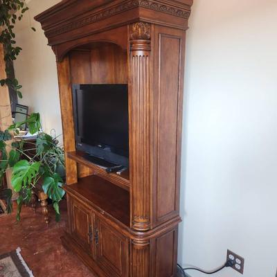 Sale Photo Thumbnail #36: A wooden entertainment armoire featuring carved details along the top and side columns. The unit includes an open space accommodating a flat-screen TV, a central shelf for media equipment, and two lower cabinet doors for additional storage. The armoire ha