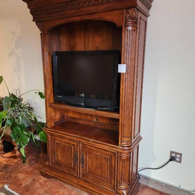 Sale Photo Thumbnail #37: A wooden entertainment armoire featuring carved details along the top and side columns. The unit includes an open space accommodating a flat-screen TV, a central shelf for media equipment, and two lower cabinet doors for additional storage. The armoire ha