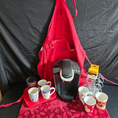 Keurig coffee maker with pod caddy - a red apron - and several Christmas themed mugs.