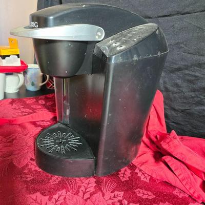 Keurig coffee maker with pod caddy - a red apron - and several Christmas themed mugs.