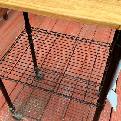 Sale Photo Thumbnail #377: It features two wire shelves and a solid wood (likely maple or butcher block) top for storage and workspace. 
The cart is designed to be functional and efficient, often used in kitchens to keep counters clutter-free. 
It includes four caster wheels which 