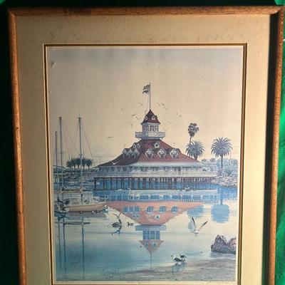 Sale Photo Thumbnail #42: A framed art print depicting a nautical cityscape featuring a historic building, boats, and palm trees reflected in calm water. The artwork is mounted within a wooden frame with a textured matte, under glass for protection. The frame is simple and complem