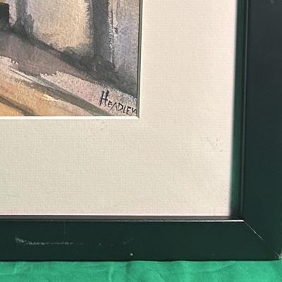 Sale Photo Thumbnail #60: Framed watercolor painting of a street scene signed by the artist, Headley, in the lower right corner. The artwork depicts a shadowed street with buildings and figures in a warm color palette. It is matted and housed in a simple black frame. Condition app
