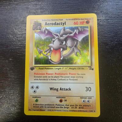 Sale Photo Thumbnail #13: First edition Aerodactyl Pokemon card from the Fossil set. The card has 60 HP and features the move Wing Attack with a damage of 30. The condition is noted as light play, indicating some wear but still collectible. This first edition card is marked with t