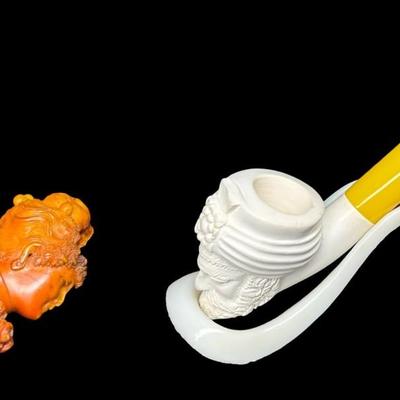 Sale Photo Thumbnail #223: Two pieces. Each is hand carved. One is unused. The amber colored piece has a crack (pictured), and may have been previously used. Two pipe stands are also included. Measurement of longest pipe: 5.75”L. [JP]