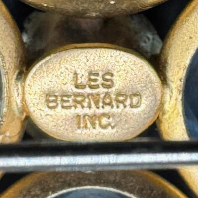 Sale Photo Thumbnail #235: This Modernist gold tone brooch is signed “Les Bernard.” It features nine green stones and four cornflower blue stones. Each stone is bezel set. Measurement: 2” x 2”. [JP]