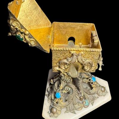 Sale Photo Thumbnail #403: This highly decorated brass piece has a marble base with ornate brass scrollwork. It contains cabochon stones and other intricate details, such as birds. This piece resembles a bird house on a pedestal. It is missing the original key. There is patina and 