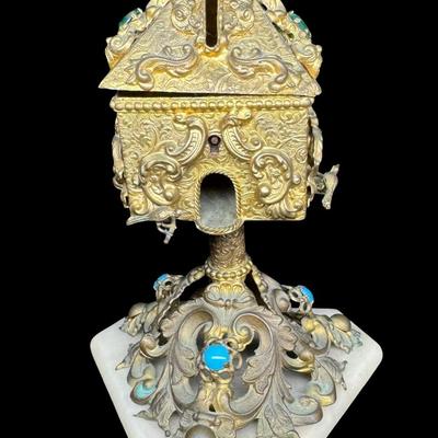 Sale Photo Thumbnail #401: This highly decorated brass piece has a marble base with ornate brass scrollwork. It contains cabochon stones and other intricate details, such as birds. This piece resembles a bird house on a pedestal. It is missing the original key. There is patina and 