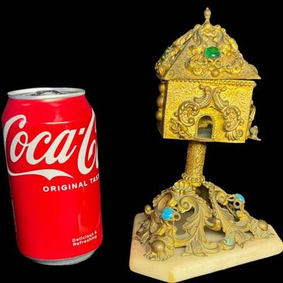 Sale Photo Thumbnail #404: This highly decorated brass piece has a marble base with ornate brass scrollwork. It contains cabochon stones and other intricate details, such as birds. This piece resembles a bird house on a pedestal. It is missing the original key. There is patina and 