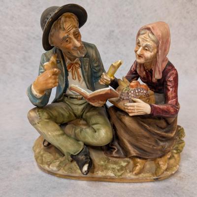 Sale Photo Thumbnail #219: Norleans Japan Porcelain Figurine 
Depicts man sitting down w/ a hat, pipe and book and a woman holding a basket 
7x8x4