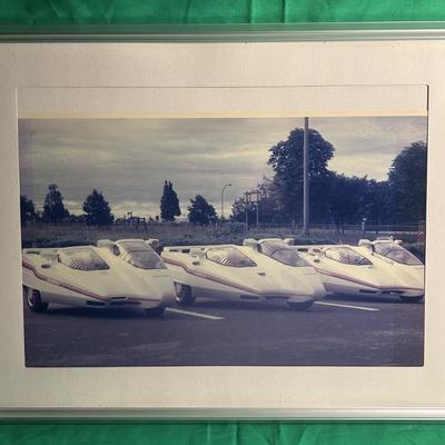 Sale Photo Thumbnail #30: A framed photograph featuring a lineup of futuristic concept cars, showcasing sleek and unique vehicle designs. The photo is encased in a metallic frame with a beige matte, and the cars are positioned in an outdoor setting with trees and a cloudy sky in t