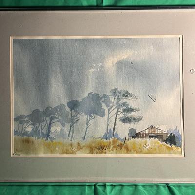 Sale Photo Thumbnail #42: A framed watercolor painting depicting a rural landscape with a row of tall trees and a small barn or farmhouse. The artwork is signed by the artist in the lower region. The painting is surrounded by a beige matte and housed in a silver-tone frame, which 