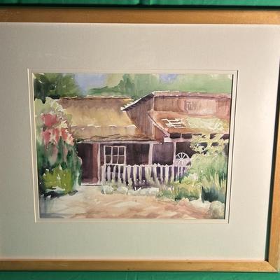 Sale Photo Thumbnail #93: Framed watercolor painting depicting a rustic house with a white picket fence, surrounded by lush greenery and flowers. The artwork is matted and presented in a gold-toned wooden frame. The painting features light and soft colors, suggesting a peaceful ru