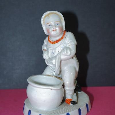 Sale Photo Thumbnail #365: Circa 1910 glazed porcelain match keep, the base of the figurine is a stike for the match, 
Little novelty of a baby about to sit on the commode. Its doll behind him. 
Height 5" 
the base measiures 4" by 3 1/4"
Good condition, slight wear to the base. and