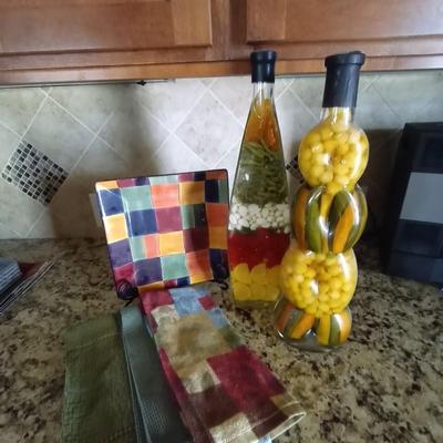 TWO GLASS BOTTLES FILLED WITH PEPPERS-DISH TOWELS AND DECO PLATE ON STAND