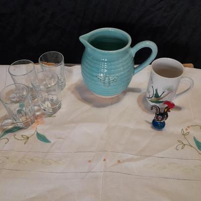 Blue pitcher with bee, coffee mug, metal rooster decor, and glasses with embroidered linen table runner