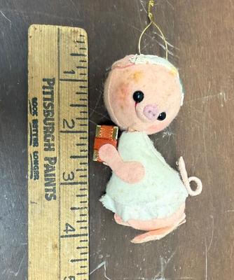Sale Photo Thumbnail #294: Analee-style pig ornament, likely vintage, featuring a round, pink face with black button eyes and a stitched snout. It has a white, layered dress with a pink patch on its arm and a curly tail. Approx. 4" tall, in good condition.