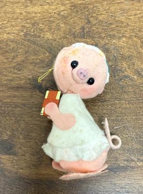 Sale Photo Thumbnail #295: Analee-style pig ornament, likely vintage, featuring a round, pink face with black button eyes and a stitched snout. It has a white, layered dress with a pink patch on its arm and a curly tail. Approx. 4" tall, in good condition.