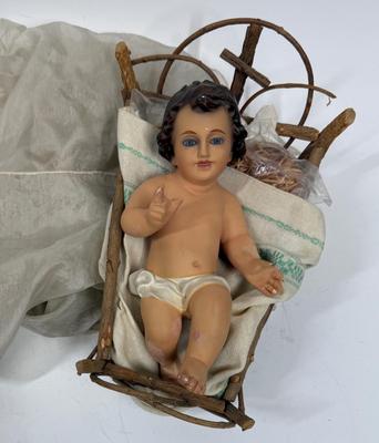Sale Photo Thumbnail #62: Recling Statue made of hand-painted porcelain with varying flesh tones. It has glass eyes and open mouth.  Cradle has straw and blanket along with a gray scarf.