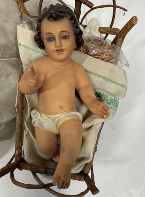 Sale Photo Thumbnail #64: Recling Statue made of hand-painted porcelain with varying flesh tones. It has glass eyes and open mouth.  Cradle has straw and blanket along with a gray scarf.
