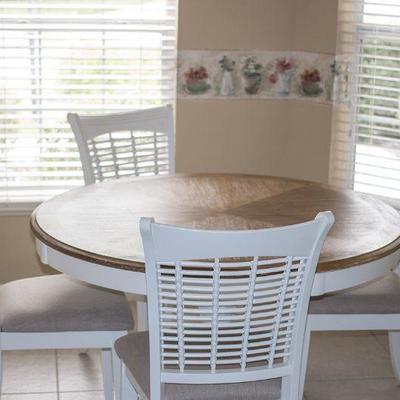 Kitchen dining set with driftwood-finish top and four white chairs with neutrally upholstered seat cushions.  Hillsdale 48" round.