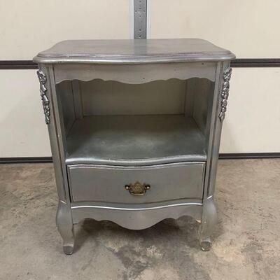 This is a bedside table or night stand with a pull out drawer and a brass handle for accent that has been painted silver. The table has a...