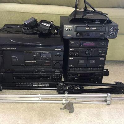 This lot contains a working Magnavox music system, a working Sanyo music system, a working Samsung VHS player, and a working NetGear WiFi...