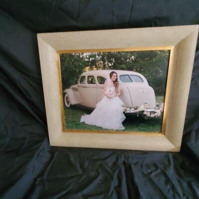 Bride captured next to antique auto.  Photo is framed in a light wooden mounting. frame size is 