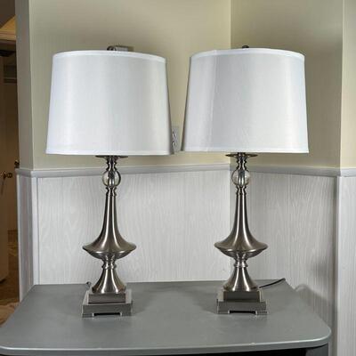 PAIR BRUSHED METAL COLUMN LAMPS | Contemporary table lamps with brushed metal and clear glass orbs, having white cylindrical shades; h....