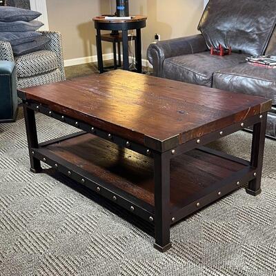 INDUSTRIAL COFFEE TABLE | Low table, having a dark wood board top and lower shelf with brass supports and tacks on a black metal block...