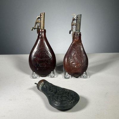 (3PC) POWDER FLASKS | Includes 2 leather black powder measures with spring loaded top, decorated with animal reliefs, and one metal...