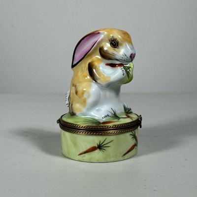 A LIMOGE RABBIT FORM BOX | A rabbit, holding a carrot on a hinged lid cylindrical box with a four leaf clover form closure