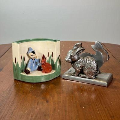 TWO NAPKIN HOLDERS | One with two bunny rabbits in metal, the other in glazed ceramic with a boy and rabbit sitting in the grass, signed...